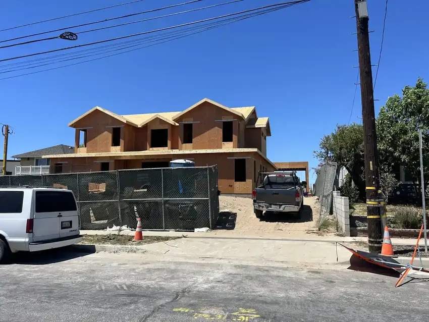 Two-story wood-framed house under construction behind site fencing, with work vehicles parked out front.