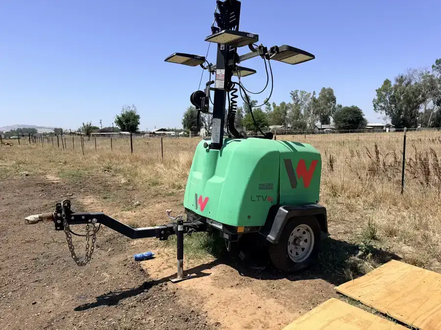 Green towable light tower with four lamp heads raised on its mast, parked on a dirt lot.